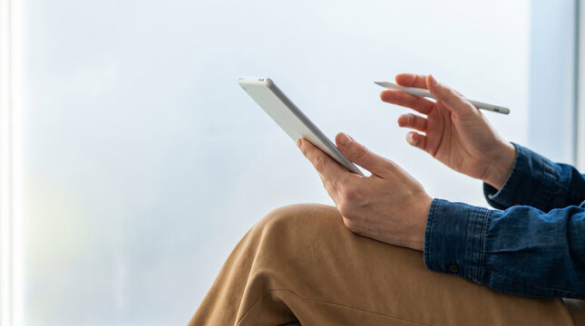 Cropped shot of person holding digital tablet and stylus pen by bright window, creative professional designer or artist working on portable device, technology and workspace concept