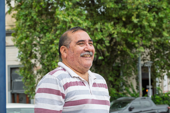 Retrato en plano americano de una persona, hombre, mayor de edad, sonriendo a c&aacute;mara, con paisaje urbano desenfocado al fondo. Imagen orientada a conceptos de bienestar, envejecimiento saludable