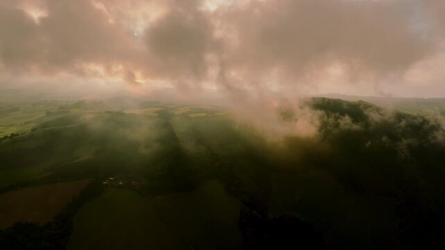 Aerial view of rolling green hills on Sao Miguel Island at sunset, with dramatic golden rays of light breaking through clouds over the misty pastoral landscape.
