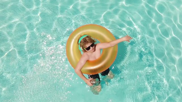 Kid stands inside orange inflatable swim ring. Kid wears headphones and dark sunglasses. Top view frames kid in turquoise pool. Bright sunlight reflects around kid in water.