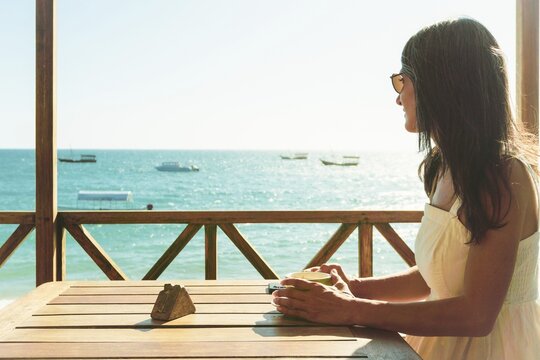 Tourist woman sitting by wooden table enjoy balcony views watch boats on seaside cafe drinking morning coffee tea, looking ocean view, Stone Town Zanzibar Tanzania travel vacation lifestyle