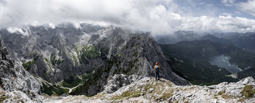 Panorama, mountaineer at the summit of the Waxenstein, view over rocky and narrow ridge of the Waxenstein ridge to Eibsee lake and H&ouml;llental with Jubil&auml;umsgrat, cloudy rocky mountain landscape, Wetterstein Mountains, Garmisch-Partenkirchen, Werdenfelser Land, Upper Bavaria, Bavaria, Germany