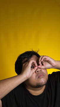 A curious young Asian male playfully forms a binocular shape with both hands over the eyes while looking upward against a bright solid yellow background. The focused person gazes away