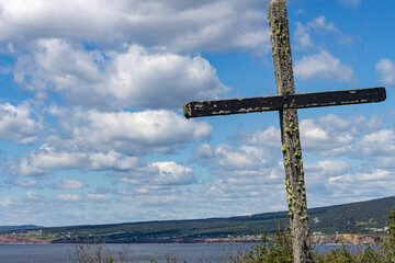 Quebec, Canada, 15 July 2026 : A weathered wooden cross covered in moss stands against a cloudy blue sky overlooking a coastal landscape. © Henry Saint John