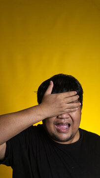 A heavily stressed young Asian male firmly covers both eyes with one hand while keeping the mouth open in a distressed groan against a vibrant solid yellow background. The deeply frustrated
