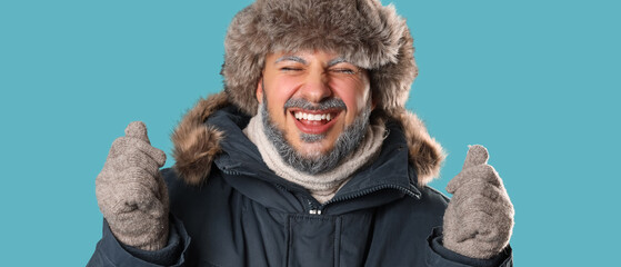 Winter joy and cold weather comfort, happy man wearing fur hat and parka with frost on beard laughing during snowy season on blue background © Pixel-Shot