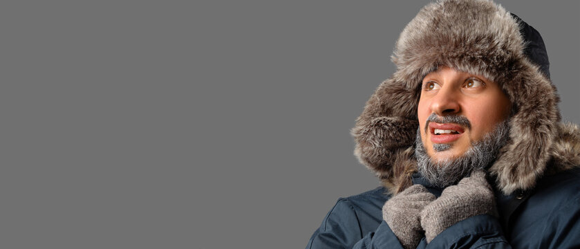 Extreme cold weather and winter survival, man wearing fur hat and parka shivering in freezing temperatures with frost on beard against gray background