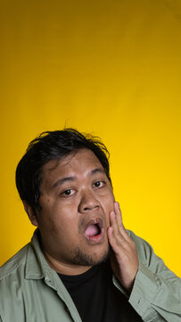 A surprised young Asian male holds one hand against the cheek with a wide open mouth against a cheerful solid yellow background. The expressive person stares directly at the lens,