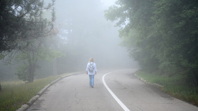 Woman walking down a foggy road into the mist alone. Back view of a female hiker with a backpack on a mountain path. Journey into the unknown and mystery concept