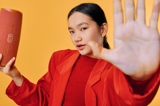 Young woman in red jacket holds portable speaker and shows stop gesture with hand in closeup for music, technology, sound and lifestyle on orange background with casual expression.