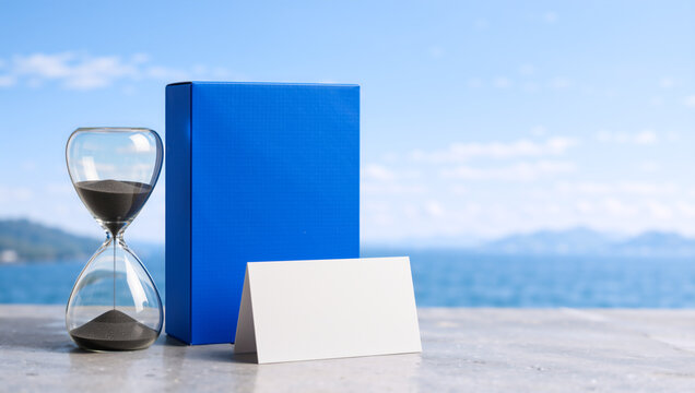 Blue box with glass hourglass and blank white card mockup by the sea. Time management and vacation planning concept on a sunny day