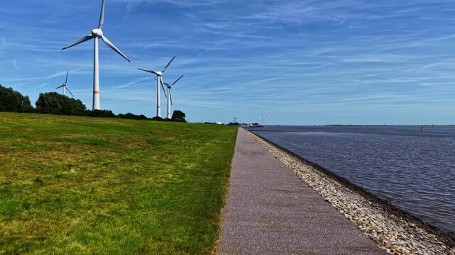 Aerial view of wind turbines standing on a green embankment along the Ems river under a clear blue sky in Emden, Niedersachsen, Germany.
