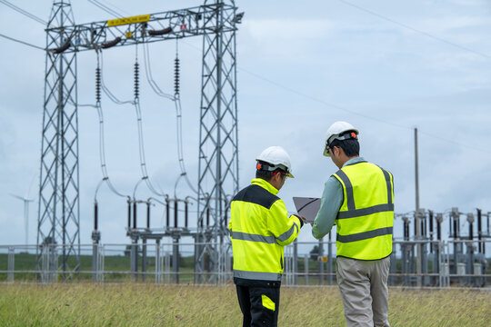 Electrical engineer and field engineer inspect high voltage substation while reviewing equipment plans during outdoor utility maintenance work under cloudy sky