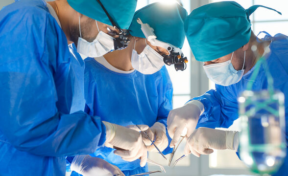 Surgeon group professional medical doctors surgical team performing serious surgery, operation, procedure on patient in surgical gown, cap, face mask, eye loupe binocular, treat injuries, diseases