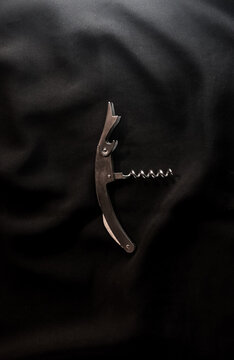 Close-up shot of a classic stainless steel waiter's corkscrew and bottle opener resting on a textured black background with dramatic lighting and soft shadows.
