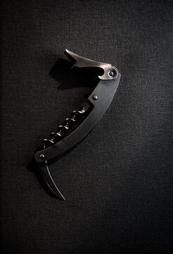 Close-up shot of a classic stainless steel waiter's corkscrew and bottle opener resting on a textured black background with dramatic lighting and soft shadows.