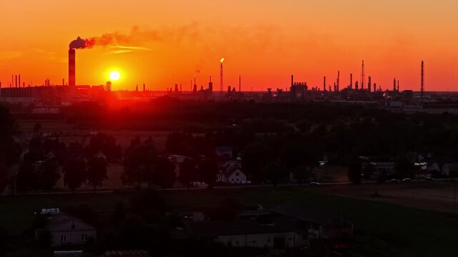 Aerial view of the ORLEN Plock Production Plant at sunset with industrial chimneys and silhouettes of refinery structures against a vibrant orange sky in Plock, Masovian Voivodeship, Poland.