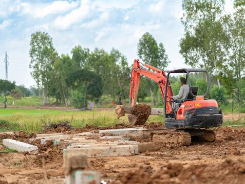 Orange mini excavator moving concrete posts on a muddy dirt terrain at an outdoor construction site.