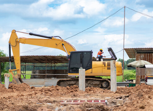 Yellow excavator machine parked at a construction site with a worker performing engine maintenance under a blue sky.