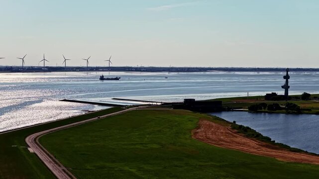 Aerial view of the Ems estuary featuring a cargo ship, wind turbines on the horizon, and a green coastal dike near the port of Emden, Niedersachsen, Germany.
