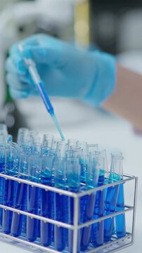 Laboratory scene showing a gloved hand using a pipette to add blue liquid sample into test tubes arranged in a rack during scientific analysis.