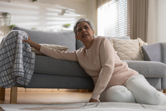 Senior safety concept. Older woman fallen beside sofa at home, reaching for support after losing balance. Senior, fall, accident, safety, elderly.