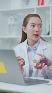 Doctor explaining cardiovascular anatomy with heart model during online medical consultation using laptop in a clinical setting.