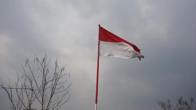 The red and white national flag of Indonesia, Sang Saka Merah Putih, fluttering from a red painted bamboo pole against a bright cloudy sky background.