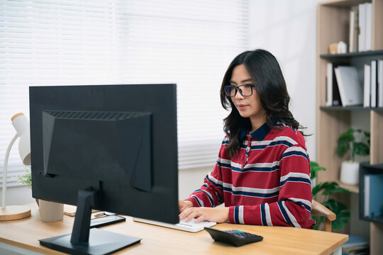 Young woman with glasses working focused on computer in modern home office with natural daylight and cozy interior