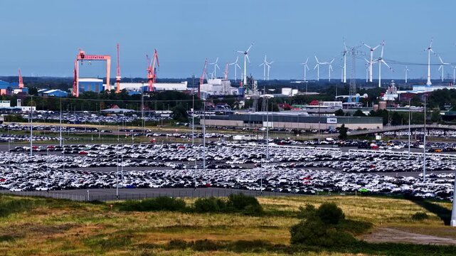 Aerial view of the Volkswagenwerk Emden car manufacturing plant, featuring rows of parked vehicles, industrial cranes, and wind turbines under a clear blue sky in Emden, Niedersachsen, Germany.