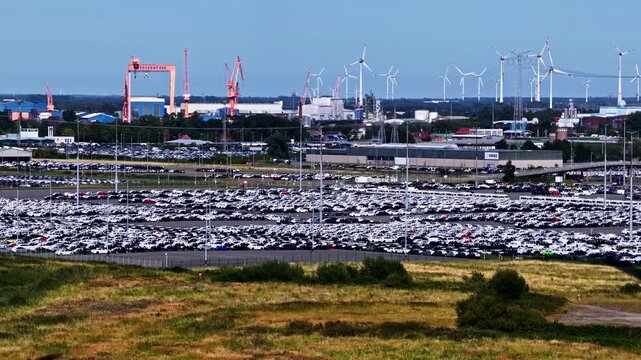 Aerial view of the Volkswagenwerk Emden car factory with rows of parked vehicles, industrial buildings, and wind turbines in the background in Emden, Niedersachsen, Germany.