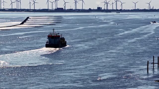 Aerial view of a cargo ship navigating the Ems river past the industrial facilities of the Volkswagenwerk Emden factory with wind turbines in the background in Emden, Niedersachsen, Germany.