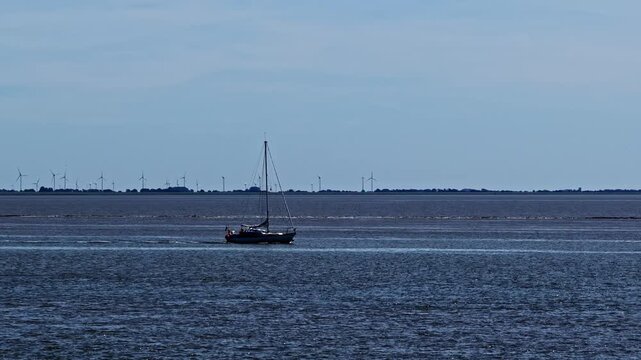 Aerial view of a lone sailboat navigating the calm waters with wind turbines visible along the distant coastline in Emden, Niedersachsen, Germany.