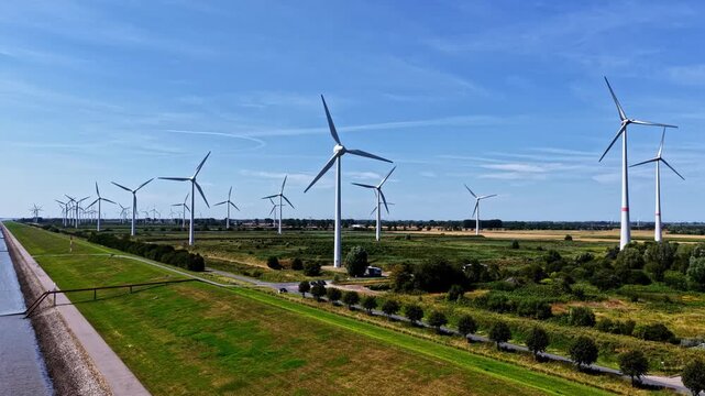 Aerial view of wind turbines standing in a green field next to a canal under a clear blue sky in Emden, Niedersachsen, Germany.
