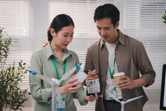 esg concept, Young Asian professionals discuss renewable energy technology while reviewing wind turbine and measuring device bright modern office, expressing focused teamwork environmental commitment