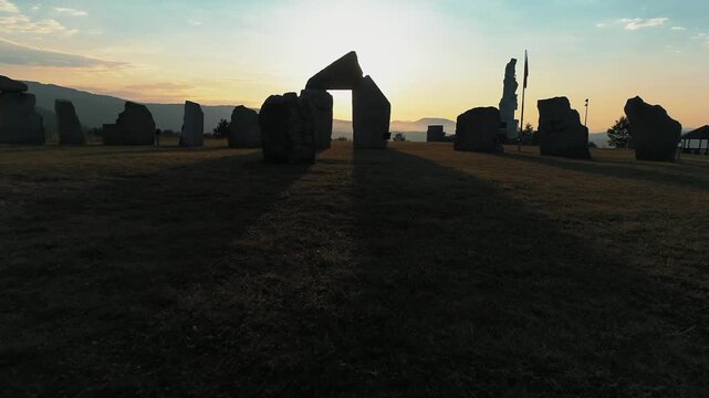 A low angle drone shot captures stonehenge replica at sunrise, sun peeking through ancient stones with monument and lake in background.