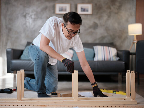 Asian mature man self-assembling wooden flat-pack furniture with DIY carpentry tools, following guide step by step, home improvement, craft installation, hobby workshop, handyman active lifestyle.
