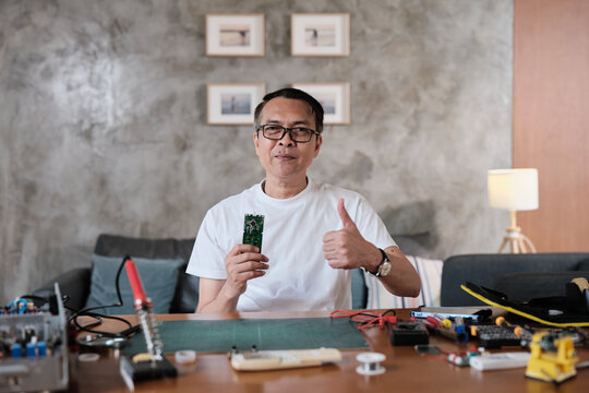 Mature Asian man showing pride in circuit board after successful soldering job, smiling, thumbs up, cheerful job achievement, home electronics appliance repair handyman, DIY fix tools, hobby workshop.