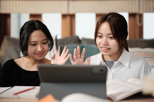 Two Asian teenage girls greeting in online class, remote learning at home using digital tablet, writing and reading textbooks, self-study for back to school, homeschooling education knowledge.