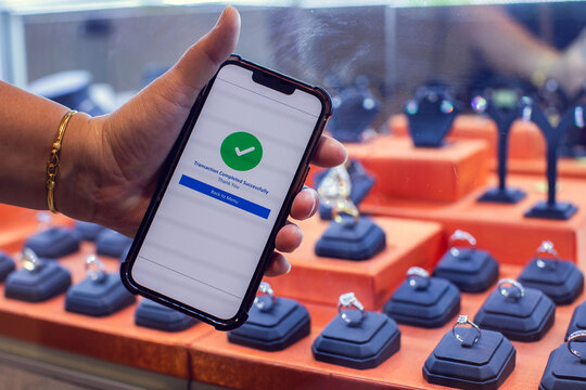Selective focus to customer hand holding a smartphone showing a simulate successful payment transaction checkmark screen inside a luxury jewelry shop with diamond rings displayed on the counter.