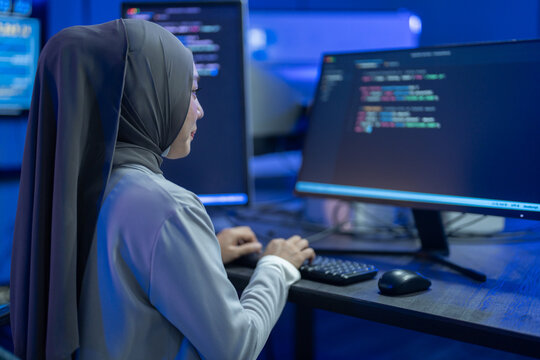 Muslim female software developer in hijab coding on multiple monitors in dark office, Happy programmer working on IT system and software development, Modern technology, cybersecurity and data concept