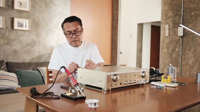 Mature Asian man disassembling and checking audio equipment, looking with magnifying glass at circuit board details for repair, home appliance DIY fix, electronics component hobby, handyman workshop.