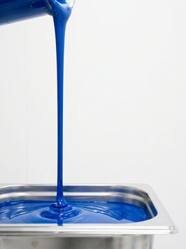 Viscous blue liquid pouring from container into stainless steel tray, demonstrating industrial material production or chemical process.