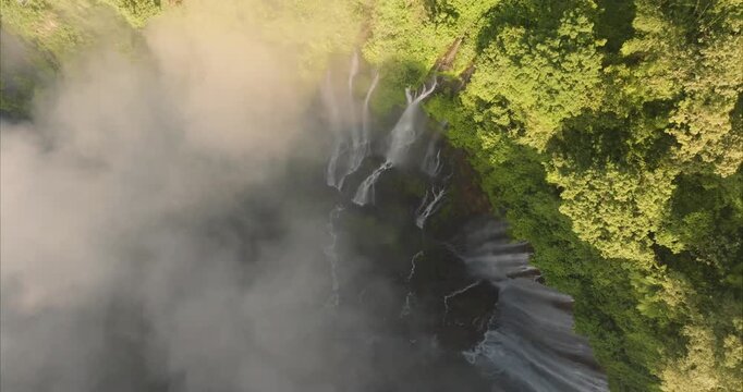 Aerial view of Tumpak Sewu Waterfall cascading down lush green cliffs into a misty gorge in Kecamatan Pronojiwo, Jawa Timur, Indonesia.