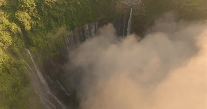 Aerial view of Tumpak Sewu Waterfall with cascading water and lush green cliffs shrouded in mist in Kecamatan Pronojiwo, Jawa Timur, Indonesia.