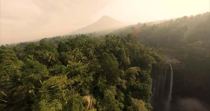 Aerial view of Tumpak Sewu Waterfall surrounded by lush tropical forest and palm trees with a mountain in the background in Kecamatan Pronojiwo, Jawa Timur, Indonesia.