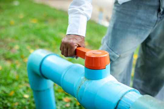 Selective focus to hand turning an orange valve handle on a blue PVC water pipe outdoors. Ideal for concepts of water saving, conservation, plumbing, utilities, and environmental control.