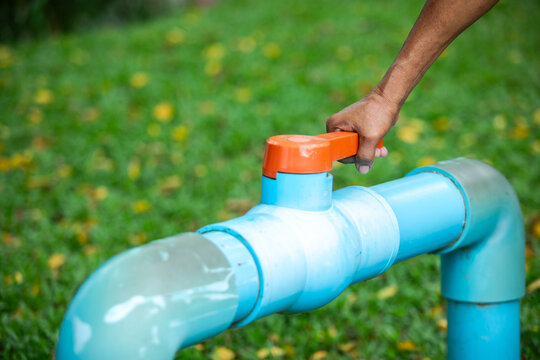 Selective focus to hand turning an orange valve handle on a blue PVC water pipe outdoors. Ideal for concepts of water saving, conservation, plumbing, utilities, and environmental control.