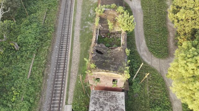 Top-down drone flyover of overgrown roofless railside brick building beside single train track and dirt road, rural Bangladesh