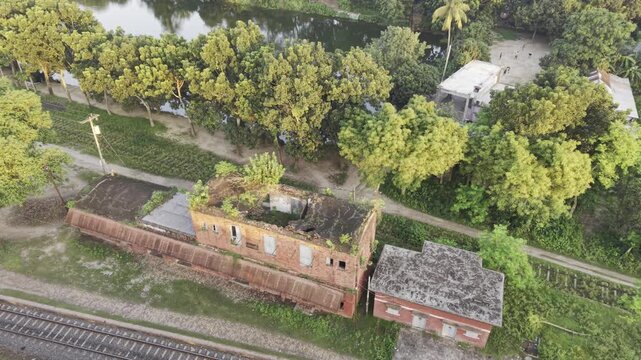 Daylight drone flyover of vegetation-filled old two-storey rail quarters by trees, canal and tracks in rural Bangladesh